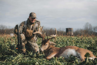 Hunter kneeling next to large buck (deer) with hunting blind in background