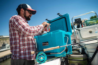 fly fishing reel and rod sitting in front of a teal grizzly 20 cooler with man opening a soda can