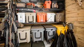 A storage room corner with metal shelving holding multiple cooler boxes and gear bags in white, gray, orange, black, and beige. A wood-paneled wall is visible on the right, along with a mounted deer skull and hanging black umbrellas.