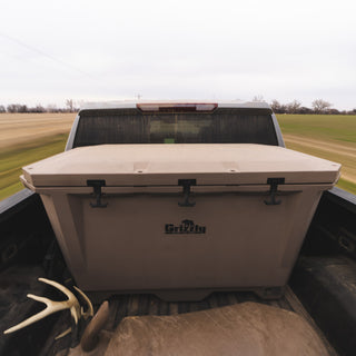The picture is showing a new Grizzly 350 cooler in the bed of a truck. The surroundings suggest that the truck is in motion as the grass and fields around it are blurred. There is also a buck decoy in the bed of the truck, in front of the G350. The large hard side cooler is tan with a black Grizzly Coolers logo on the front.