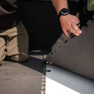 The image is from the inside of the Alpha Hunting Blind displaying a closeup of the flooring. A man is kneeling down and has pulled back a piece of the flooring which is notched and connects seamlessly to the other floor insert. The unique hunting blind flooring is far superior to other materials such as wood floors. It is weather-resistant, silent, provides comfort under foot, and quiet. The floor color is a very dark charcoal color and fits snugly in the Alpha Hunting Blind.