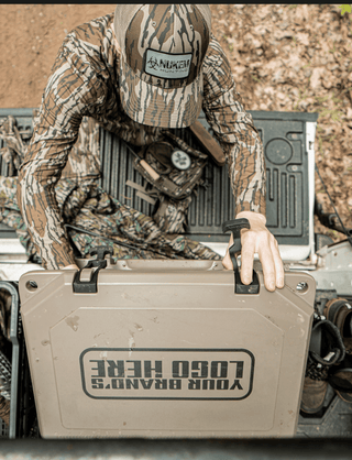The image is an aerial view of the lid of a Grizzly cooler that someone is unlatching and opening. On top of the lid, you can see an imprint that reads "your brand's logo here". Grizzly Coolers offers custom branding on their products including custom branded coolers and custom branded drinkware. The hard sided cooler in this image is tan and is in a truck bed with the tailgate down. There's a turkey decoy and hunting boots in the image on each side of the cooler.