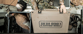 The image is an aerial view of the lid of a Grizzly cooler that someone is unlatching and opening. On top of the lid, you can see an imprint that reads "your brand's logo here". Grizzly Coolers offers custom branding on their products including custom branded coolers and custom branded drinkware. The hard sided cooler in this image is tan and is in a truck bed with the tailgate down. There's a turkey decoy and hunting boots in the image on each side of the cooler.