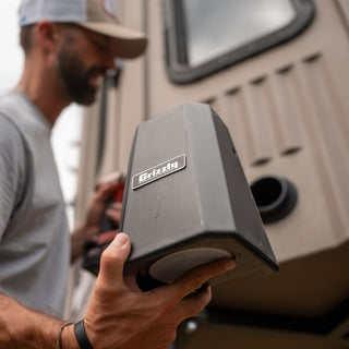 The picture is a man standing outside of an Alpha Hunting Blind holding the Power Vent in his right hand. He is holding a drill in his left hand. The man is installing the Power Vent in the elevated hunting blind. The Power Vent circulates fresh air and eliminates human scent so your prey cannot smell your presence regardless of wind direction.