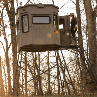 The image shows a woman at the top of the tower kit ladder entering a Grizzly Alpha hunting blind. The blind is whitetail color, a brown shade that blends in seamlessly with the wooded surroundings. The hunting blind has vertical and horizontal glass windows, and you can see a blackout shade in use on the door.